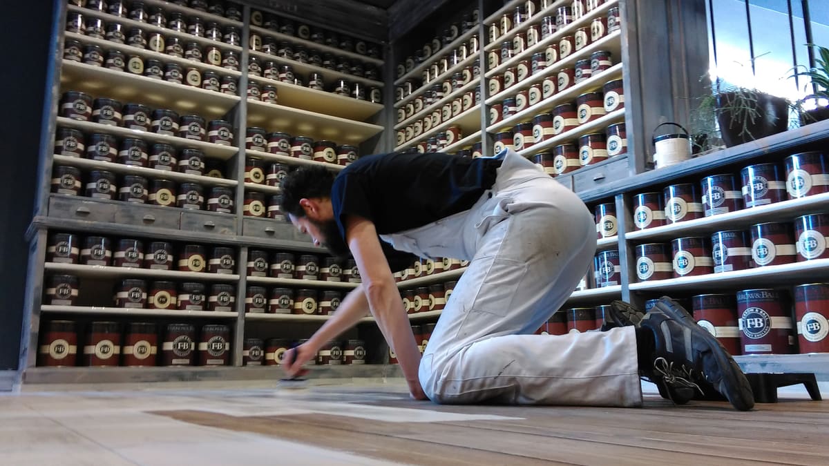 Een schilder brengt op zijn knieën Pro Finish Krijtverf aan op een houten vloer, voor een kastwand vol blikken Farrow & Ball